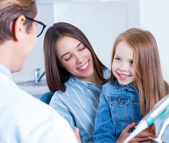 Young girl smiling at her dentist while sitting in her mothers lap