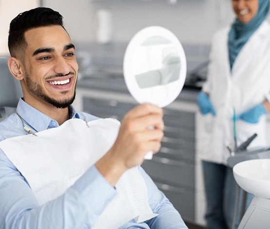 Man in dental chair smiling at his reflection in mirror with dentist blurry in background