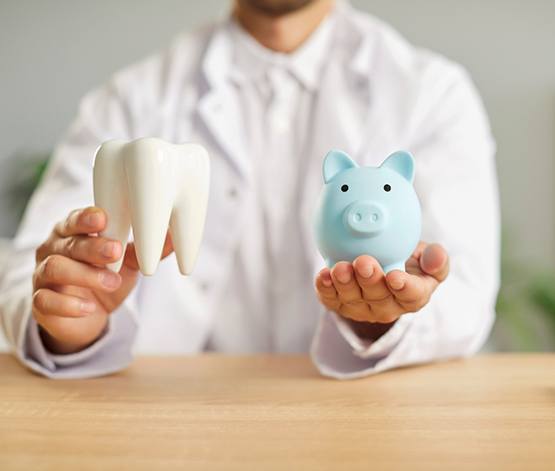 Man in white dentist jacket at desktop holding model tooth and blue piggy bank